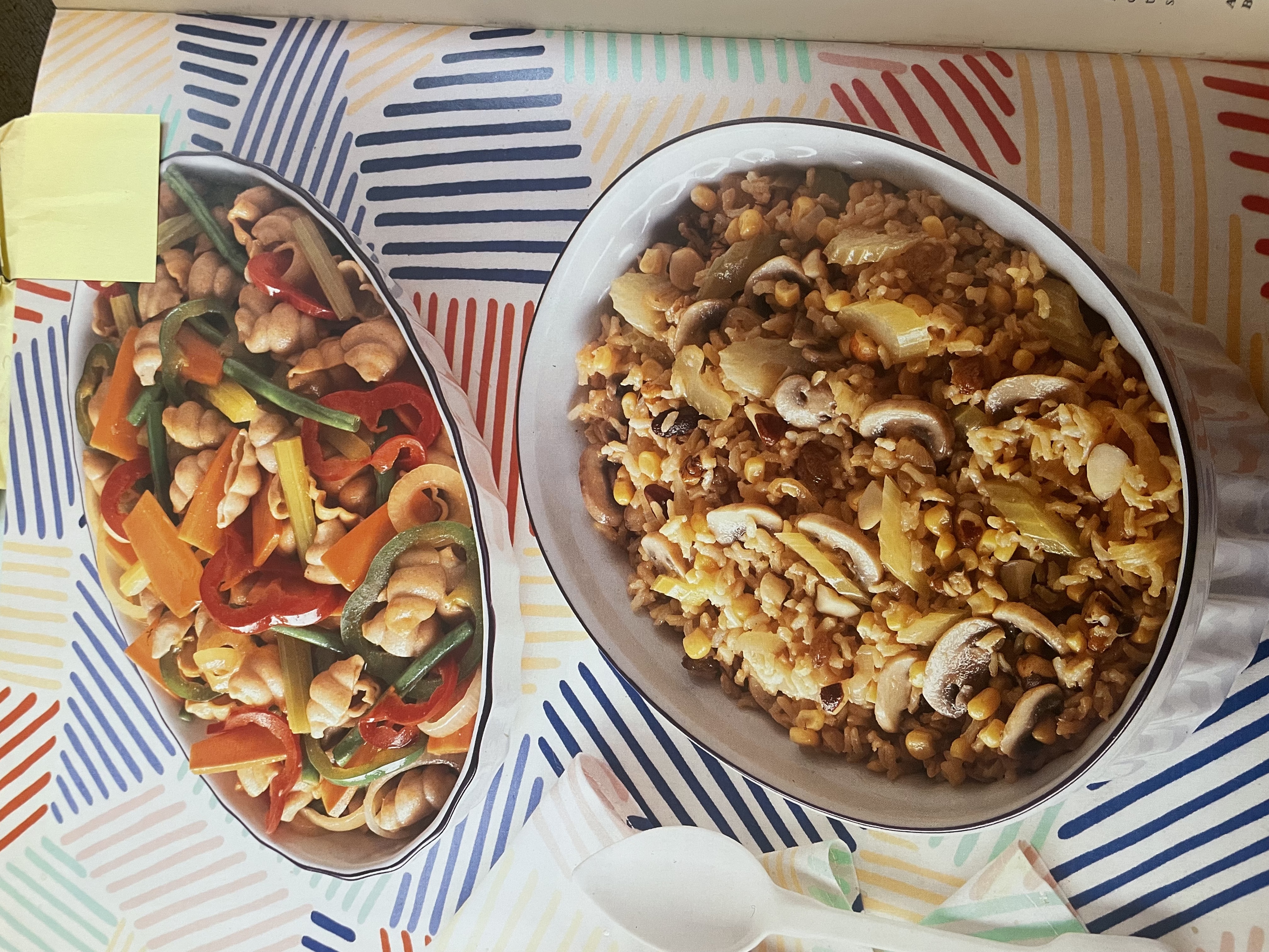 Two serving dishes of food, a fried rice and a pasta salad, on top of a table cloth with primary colored blocks of parallel lines in a random arrangement